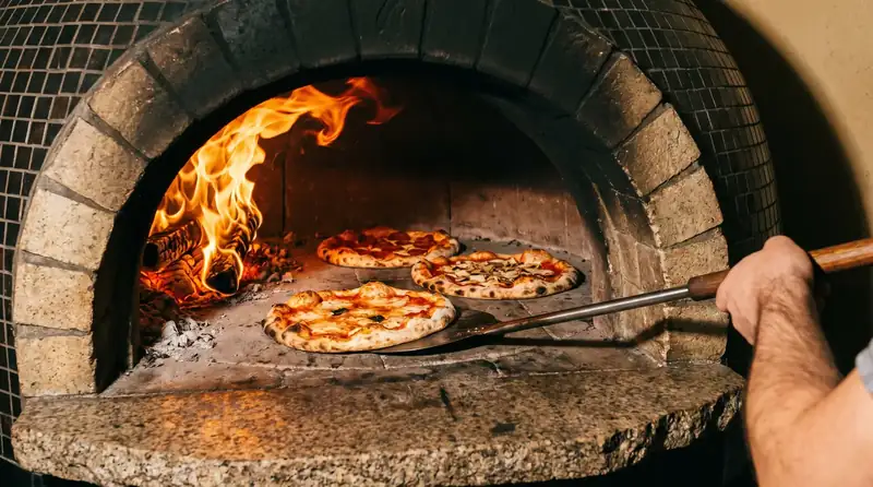 Boca do forno a lenha com chamas e três pizzas assando no piso de pedra